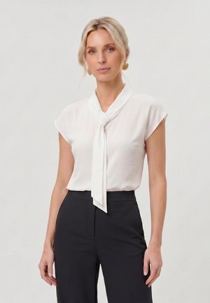 Blond hajú nő arany virágos fülbevalóval, fehér rövid ujjú blúzzal nyakkendővel és fekete, magas derekú nadrággal, egyszerű háttér előtt áll.