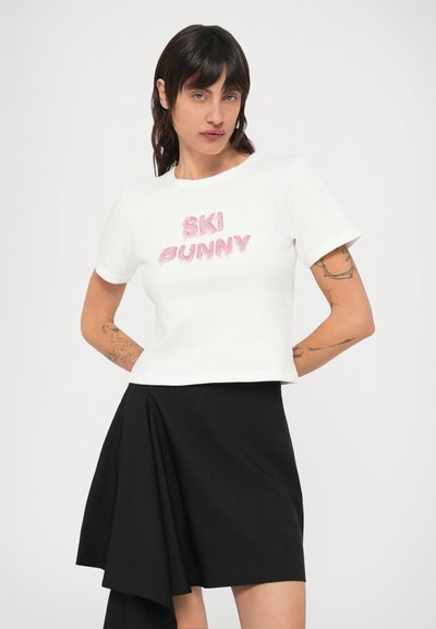 Biele tričko s krátkym rukávom a ružovým nadýchaným nápisom "SKI BUNNY", doplnené čiernou asymetrickou sukňou s záhybovou látkou.