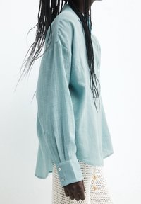 Chemise oversized bleu clair à manches longues en tissu texturé, dotée de poignets à boutons et d'une coupe décontractée, portée avec un bas en crochet.
