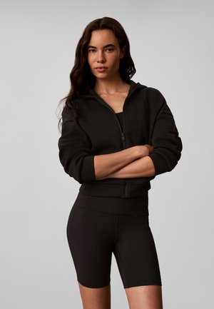 Vrouw met lang donker haar draagt een zwarte zip-up hoodie en zwarte biker shorts, staat met gekruiste armen tegen een effen grijze achtergrond.