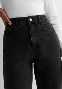 Person som bär högmidjade svarta denimjeans med knapp framtill och en vit ribbad långärmad topp, med handen vilande på höften.