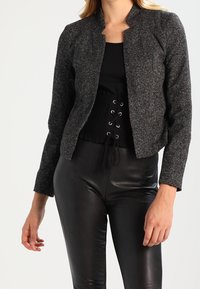 Femme portant une veste texturée gris foncé, un corset noir à lacets et un pantalon en cuir noir, debout devant un fond blanc.