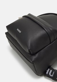 Mochila de cuero negro con acabado texturizado, que cuenta con dos compartimentos con cremallera y una correa para el hombro con el logo "HUGO" en blanco.