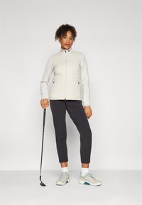 Ljust quiltat västar med hög krage, grå långärmade tröja, kombinerat med mörka byxor och beige sneakers. Inkluderar ett golfklubbs-tillbehör.