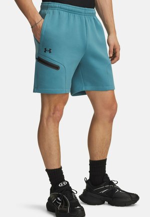 Turkooizen sportshorts met een elastische tailleband, een zwarte zijritszak en vlakke, omgezette zoom. Draagt zwarte sneakers en sokken.