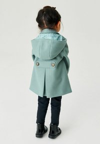 Manteau en laine vert clair avec capuche, doublure bleue à motifs et deux boutons marron à l'arrière, porté avec un jean noir et des bottes brillantes.