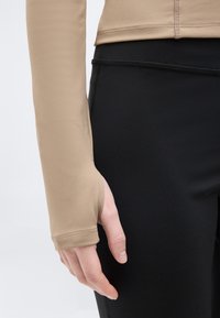 Μπεζ μπλούζα με μακριά μανίκια και τρύπα για τον αντίχειρα, φορεμένη με μαύρο εφαρμοστό κολάν, με κοντινό πλάνο του αριστερού χεριού και του χεριού να ακουμπούν δίπλα στο πόδι.