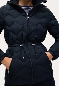 Námořnická modrá prošívaná bunda s úzkým pasem, která má plný zip, kapuci na šňůrku a boční kapsy se stříbrnými detaily.