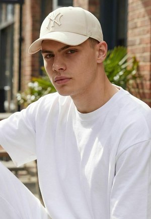 Junger Mann mit beiger New York Yankees-Kappe und einfachem weißen T-Shirt, sitzt draußen vor unscharfem Pflanzen- und Ziegelwandhintergrund.