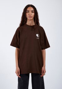Camiseta oversized marrón de algodón, con un gráfico de corazón blanco en el lado izquierdo del pecho. La camiseta tiene mangas cortas y un corte holgado.