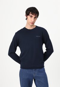 Marineblå langermet skjorte laget av bomull, med rund hals og en subtil logo på brystet, kombinert med blå denim jeans.