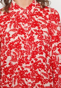 Camisa de botão vermelha e branca com um padrão floral, tecido leve e colarinho clássico. Os botões são brancos e estão espaçados uniformemente.