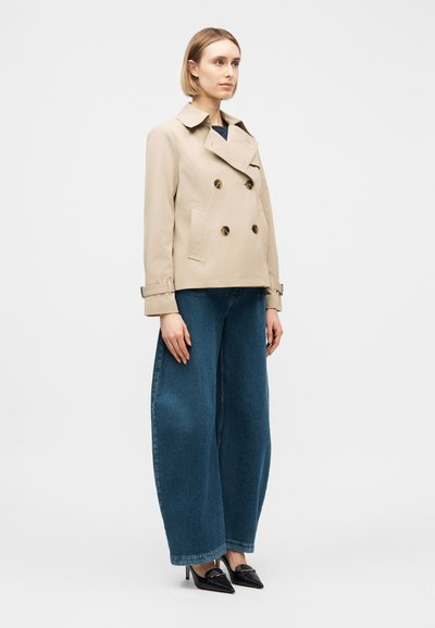 Femme debout portant une veste beige croisée, un jean bleu à jambes larges et des talons noirs pointus, sur un fond clair uni.