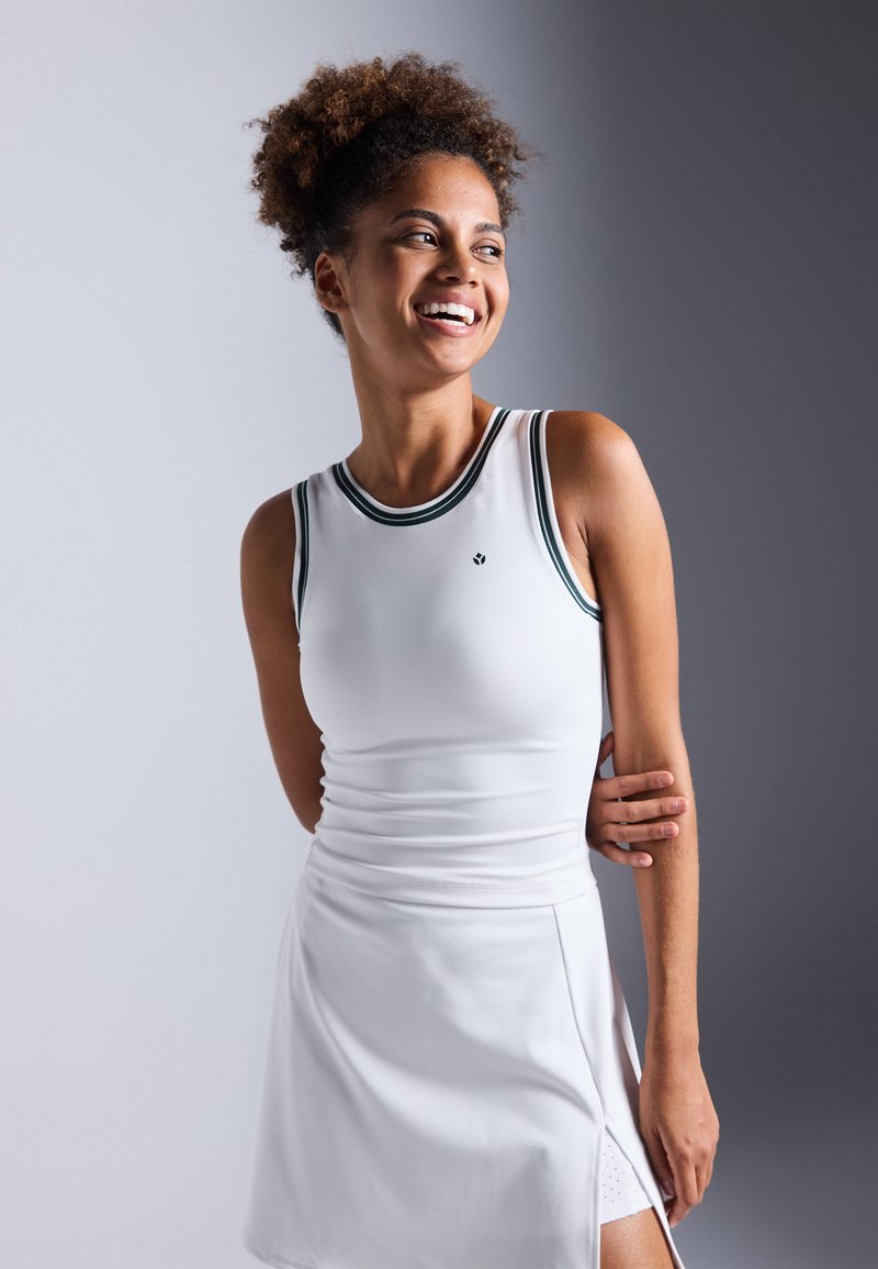 Haut de sport sans manches blanc avec des accents vert foncé, au design ajusté, et un détail froncé sur le côté, associé à une jupe blanche assortie.