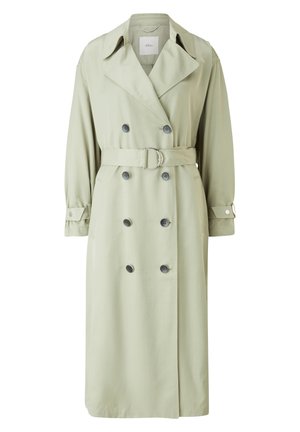 Femme portant un trench-coat croisé vert clair avec ceinture, pantalon blanc à jambes larges et sandales beiges à talons ouverts.