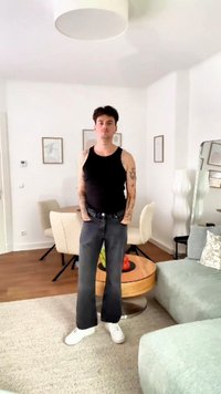 Svart tank topp, grå vide jeans, hvite sneakers. Tatoveringer synlige på armene. Moderne interiør med lyse møbler og dekorative detaljer.