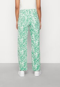 Calças verdes e brancas com um padrão solto, apresentando um design abstrato, cintura alta e bolsos laterais. Usadas com ténis brancos.