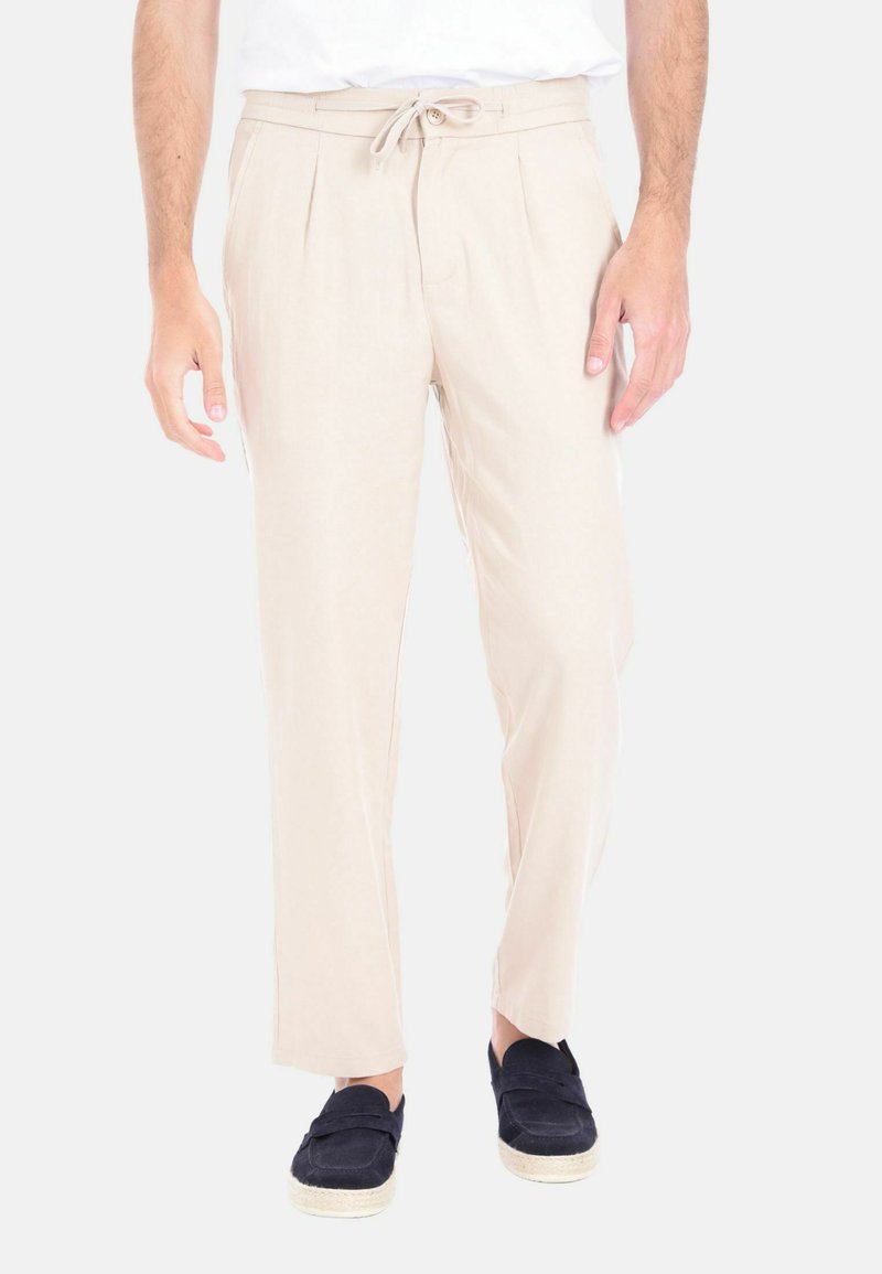 Pantaloni beige leggeri con vita regolabile, con pieghe frontali e gamba dritta. Indossati con scarpe scure senza lacci. Texture liscia.