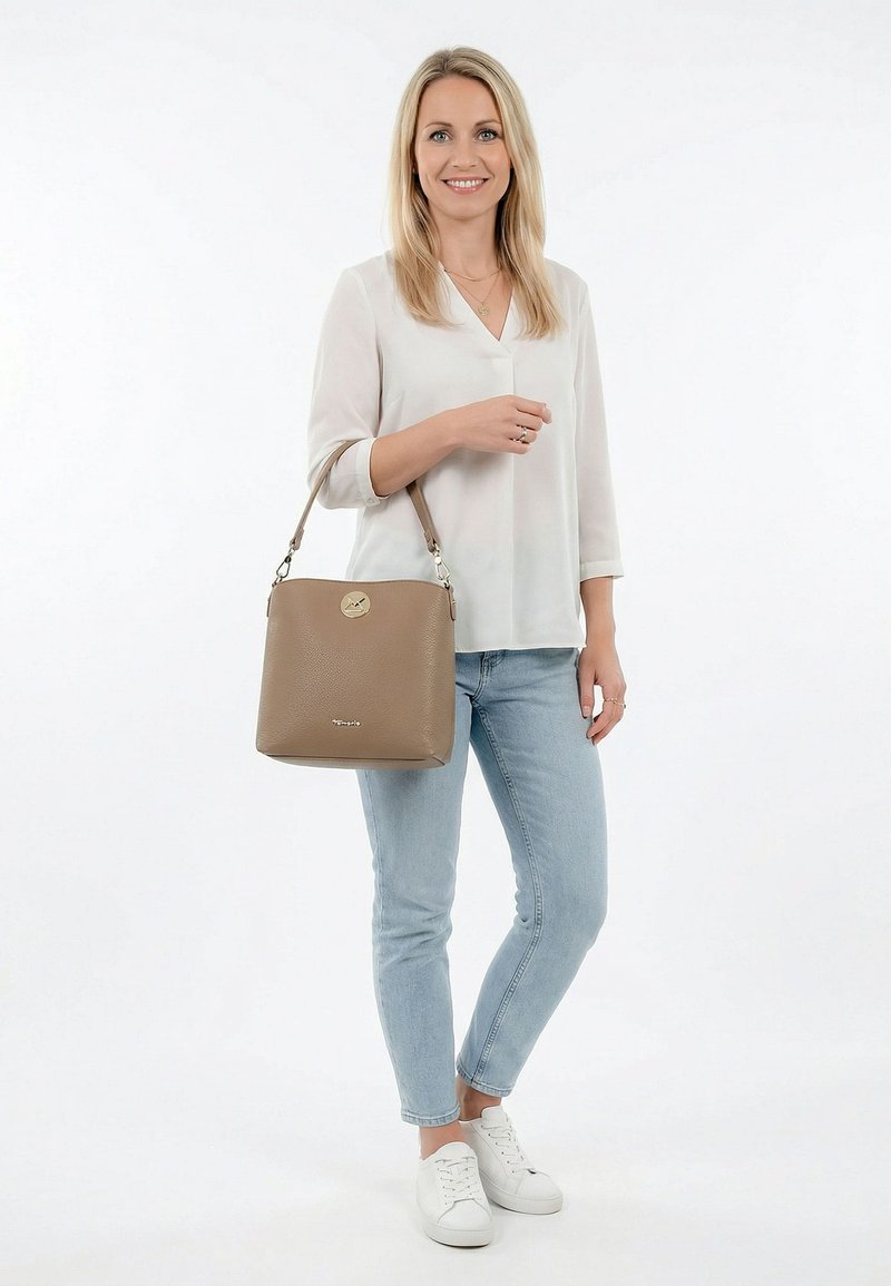 Woman in white blouse and light blue jeans carrying a taupe handbag, standing and smiling against a plain white background.