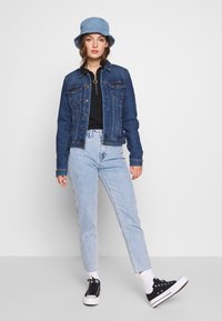 Denim jacket, dark blue, with button closure and two front pockets. Paired with light blue jeans, black sneakers, and a light blue bucket hat.
