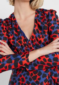 Klänning i omslag med en djärv röd och blå leopardtryck på ett glatt tyg. Djup V-halsringning och långa ärmar med subtila samlingar vid axlarna.