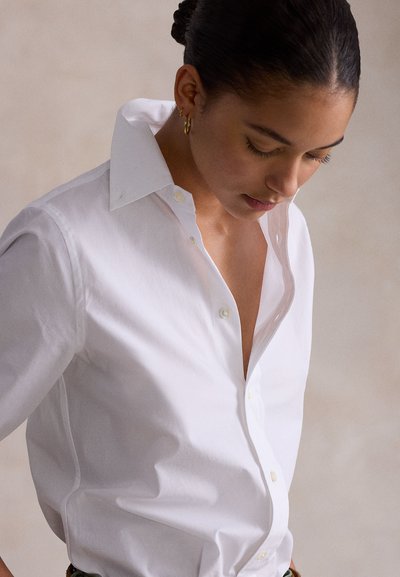 Γυναίκα με σκούρα μαλλιά σε sleek κότσο, φορά χρυσούς κρίκους και λευκό πουκάμισο με κουμπιά, κοιτάζοντας προς τα κάτω σε ουδέτερο φόντο.