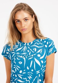 Camisa de mangas cortas azul con un patrón de hojas blancas, escote redondo y detalle de logo en el frente, fabricada con una tela texturizada.