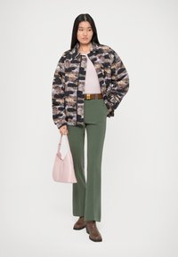 Jachetă căptușită de camuflaj în negru, maro și bej; top roz deschis; pantaloni evazați verzi; cizme cu toc maro; geantă roz.