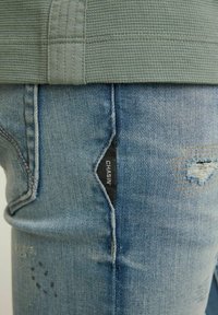 Gros plan sur un jean en denim bleu avec des détails usés et une petite étiquette noire en tissu portant l'inscription « CHASIN' » près de la hanche, porté avec un haut vert texturé.