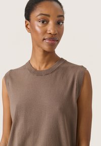 Frau mit kurzen Haaren, die ein ärmelloses, taupefarbenes Stricktop trägt, steht mit neutralem Gesichtsausdruck vor einem schlichten, hellen Hintergrund.