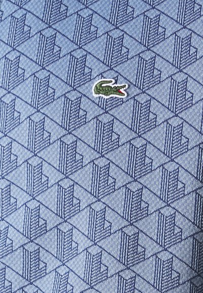 Tissu bleu à motifs avec un motif géométrique et un petit logo de crocodile vert. La texture semble lisse et tissée.
