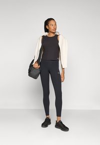 Top preto sem mangas e leggings justas com uma textura suave, acompanhados de um hoodie creme caído sobre os ombros e sapatilhas desportivas pretas.