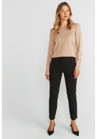 Beige pitkähihaisen neuleen parina slim-fit mustat housut ja mustat korkokengät, neutraalilla taustalla.