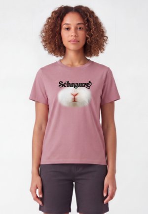 Frau mit lockigem Haar, die ein pinkes T-Shirt trägt und eine Nahaufnahme von der Nase und dem Mund eines weißen Kaninchens zeigt, mit dem Wort "Schnauze" darüber.