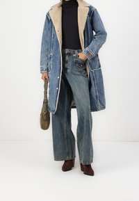 Manteau en denim avec un col en shearling beige, associé à un jean taille haute à pattes d'éléphant. Des bottes à talons marron et un sac vert complètent le look.