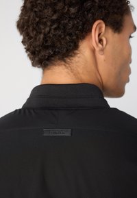 Melna polo krekla ar strukturētu apkakli un Calvin Klein etiķeti aizmugurē. Gluda materiāla un pielāgota piegriezuma, demonstrējot minimālisma dizainu.