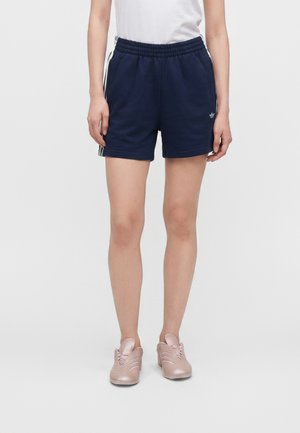 Person iført marineblå atletikshorts med hvide striber, en hvid skjorte og lyserøde sneakers, stående mod en ensfarvet baggrund.