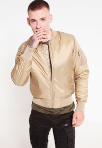 Chaqueta bomber beige hecha de tejido brillante, con cerradura de cremallera al frente, puños acanalados y un bolsillo de parche en la manga izquierda.