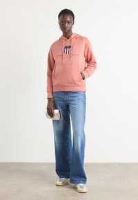 Felpa rosa con logo e design 1949, abbinata a jeans a gamba larga blu chiaro e sneakers argento. Il modello tiene una clutch trasparente.