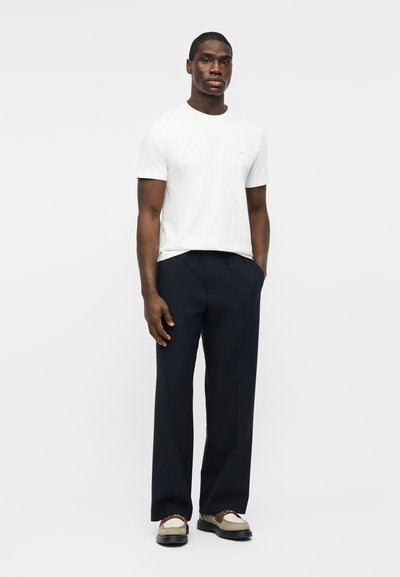 Άνδρας που στέκεται φορώντας λευκό πουκάμισο με κοντά μανίκια, σκούρο φαρδύ παντελόνι και δίχρωμα μοκασίνια, μπροστά σε απλό γκρι φόντο.