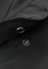 Tessuto nero con una leggera lucentezza, caratterizzato da due bottoni a pressione in metallo. La texture appare liscia con cuciture visibili lungo i bordi.