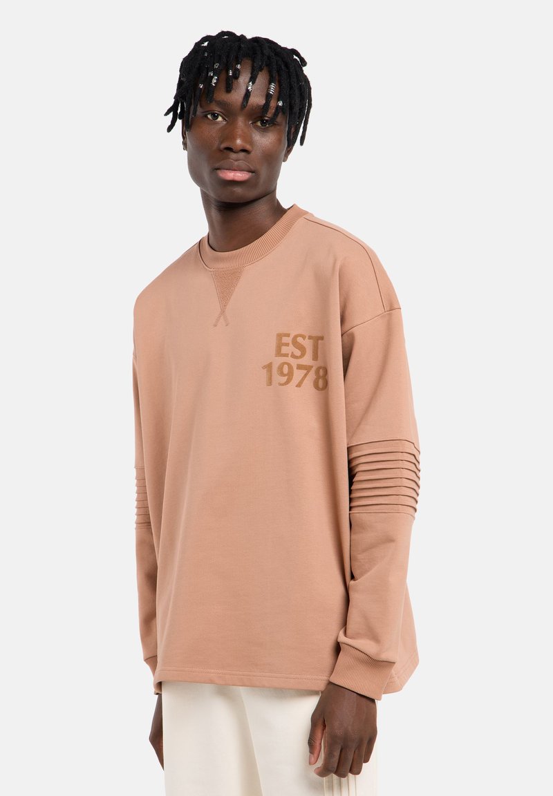 Sweat-shirt rose clair avec des accents côtelés sur les manches et "EST 1978" imprimé en marron. Présente un col rond et une coupe décontractée. Matière en mélange de coton.