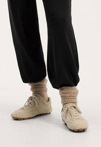 Beige suede tennarit, joissa on pyöreä kärki, nauhat ja kumipohjat; yhdistetty vaaleanruskeisiin ribbipolkimiin mustien rentojen housujen kanssa.