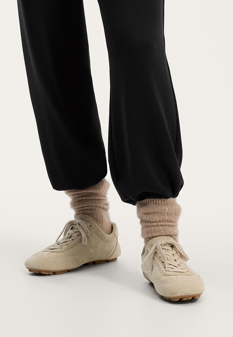 Beige suede tennarit, joissa on pyöreä kärki, nauhat ja kumipohjat; yhdistetty vaaleanruskeisiin ribbipolkimiin mustien rentojen housujen kanssa.