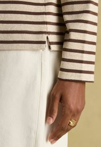 Braun gestreiftes, geripptes Stricktop mit einem seitlichen Schlitz, getragen über einem weißen strukturierten Stoffunterteil. Eine Hand mit einem goldenen Ring ist sichtbar.
