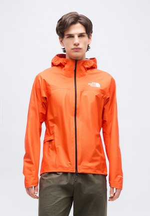 Νεαρός άνδρας που φοράει μια φωτεινή πορτοκαλί ζακέτα με κουκούλα, φερμουάρ μπροστά και τσέπες, στέκεται μπροστά σε ένα απλό φωτεινό φόντο.