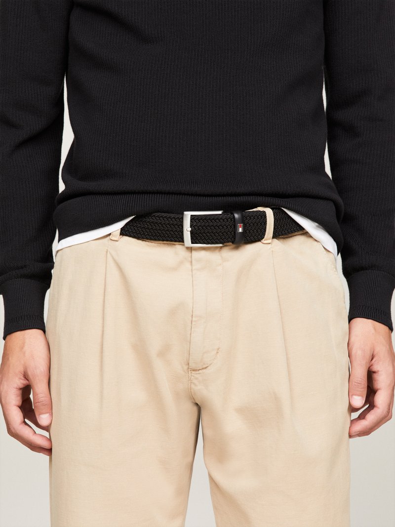Mann in beigen, plissierten Hosen mit einem schwarzen, geflochtenen Gürtel und einem schwarzen Langarmpullover, die Hände entspannt an den Seiten.