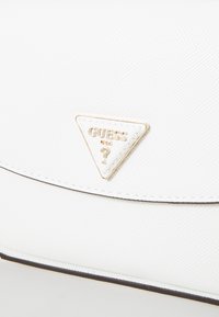 Borsa a mano bianca con una finitura testurizzata, caratterizzata da un logo hardware triangolare dorato inciso con "Guess USA". Chiusura con bordo foderato e cuciture visibili.