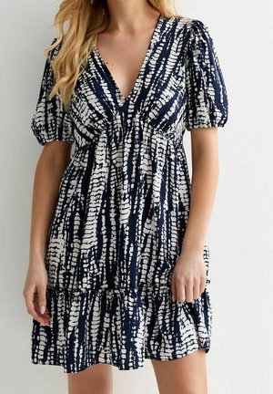Kobieta w krótkiej granatowej sukience z białymi, abstrakcyjnymi pionowymi paskami, z bufkami na krótkich rękawach i dekoltem w serek.