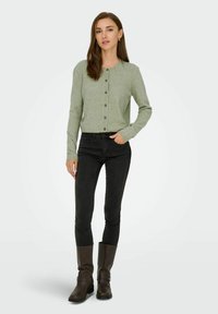 Gaiši zaļš kardigāns ar priekšējiem pogām, apvienots ar melniem šauriem džinsiem un brūnām līdz ceļgaliem augstām zābakiem. Gluda tekstūra, ikdienišķs dizains.
